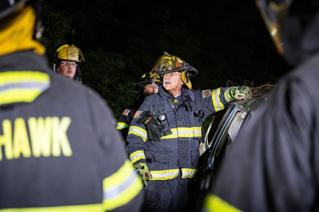 A firefighter in full gear stands beside a damaged vehicle at night, speaking to other firefighters who are gathered around, their reflective uniforms illuminated in the dark.