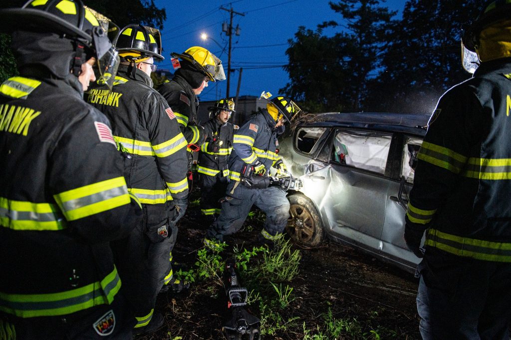 Firefighters in full gear gather around a damaged vehicle at night, working to respond to an emergency situation. The surrounding area is dark, illuminated by their equipment and vehicle lights.