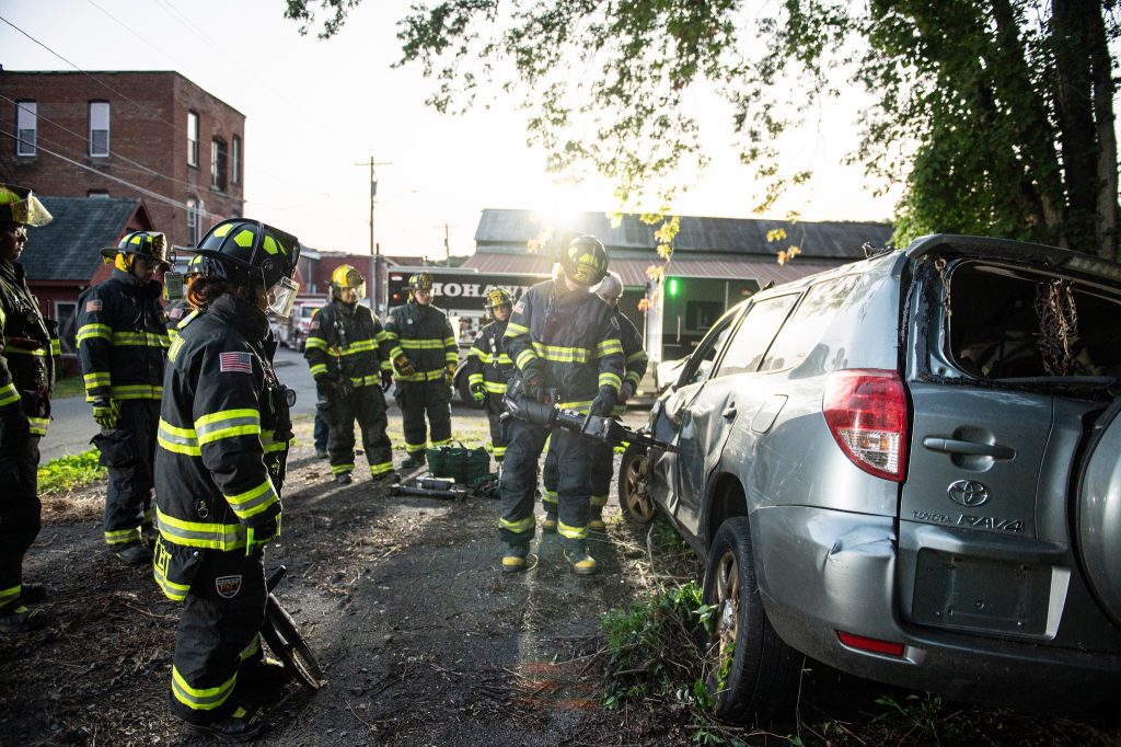A group of firefighters in full gear stand around a damaged Toyota RAV4 with broken windows, assessing the scene in daylight, with trees and buildings visible in the background.