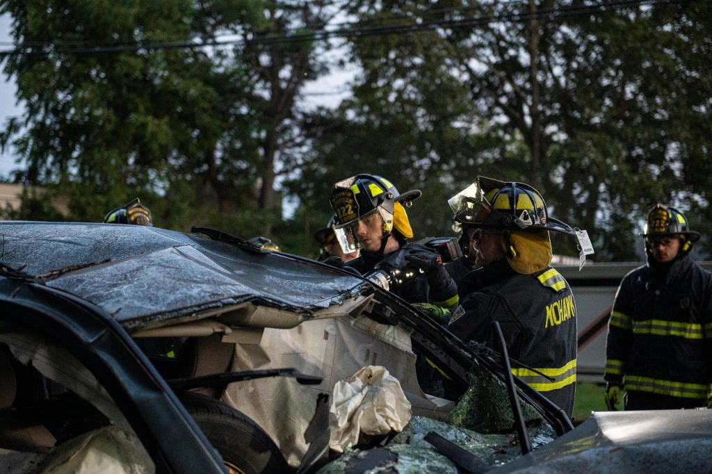 Firefighters in full gear use tools to cut through the roof of a severely damaged car during a rescue operation, surrounded by trees and power lines.