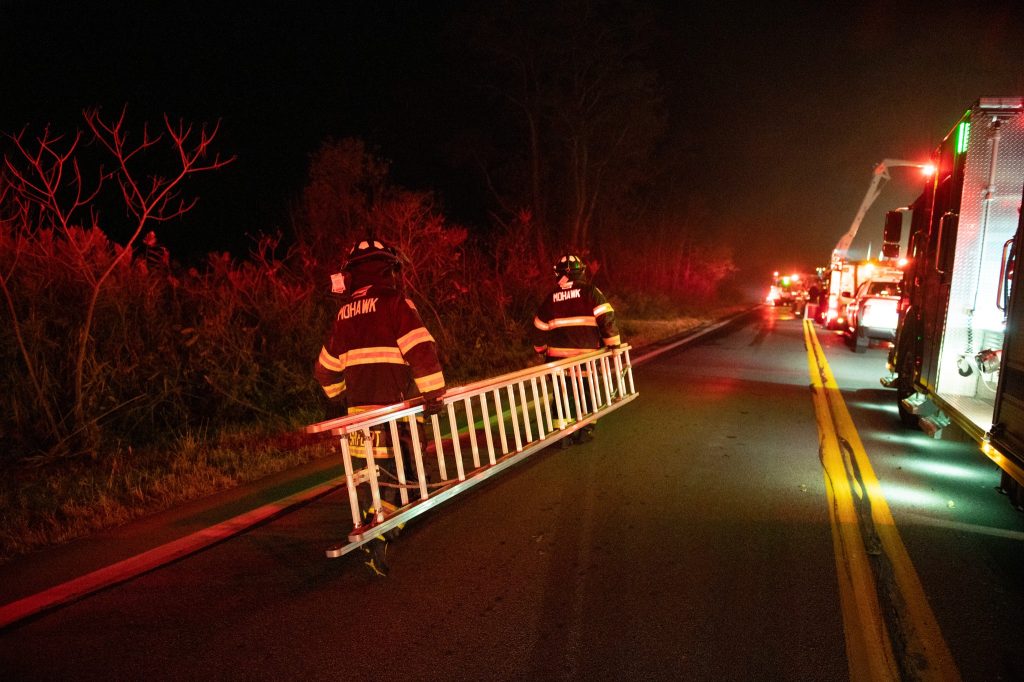 Two firefighters in gear carry a long ladder down a road at night, illuminated by red emergency lights from fire trucks parked nearby. Trees and bushes line the side of the road.