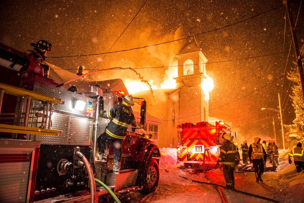 Firefighters in winter gear battle a large blaze engulfing a church at night. Flames and smoke pour from the building’s steeple. Fire trucks and hoses are visible in the snowy, illuminated scene.