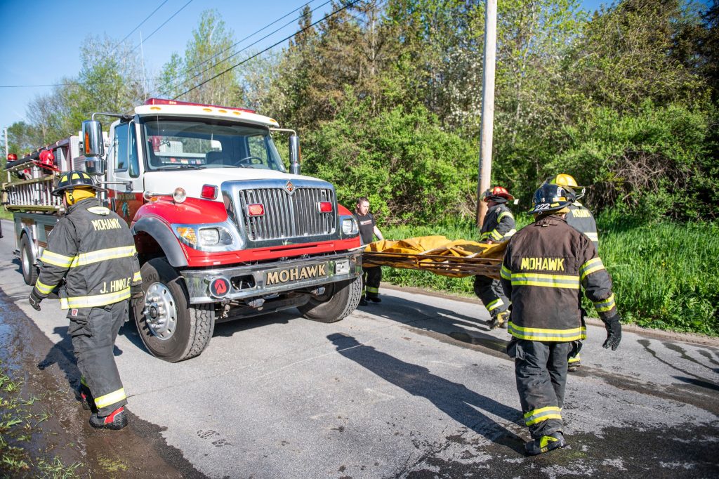 Four firefighters in Mohawk uniforms carry a stretcher near a red fire truck parked on a road, with green trees and utility lines in the background.